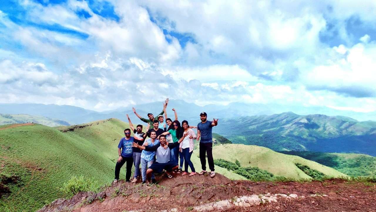 A group of trekkers cheering on a grassy ridge in the Western Ghats with green hills and clouds behind them