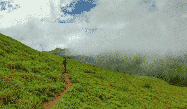 A trekker walking a narrow dirt trail across a misty grassland slope in the Western Ghats